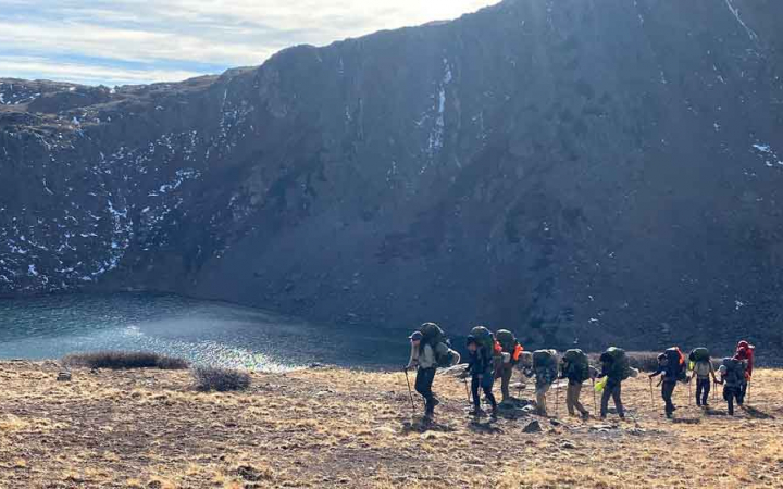 A group of people wearing backpacks hike in a line in front of a body of water in a mountainous landscape.